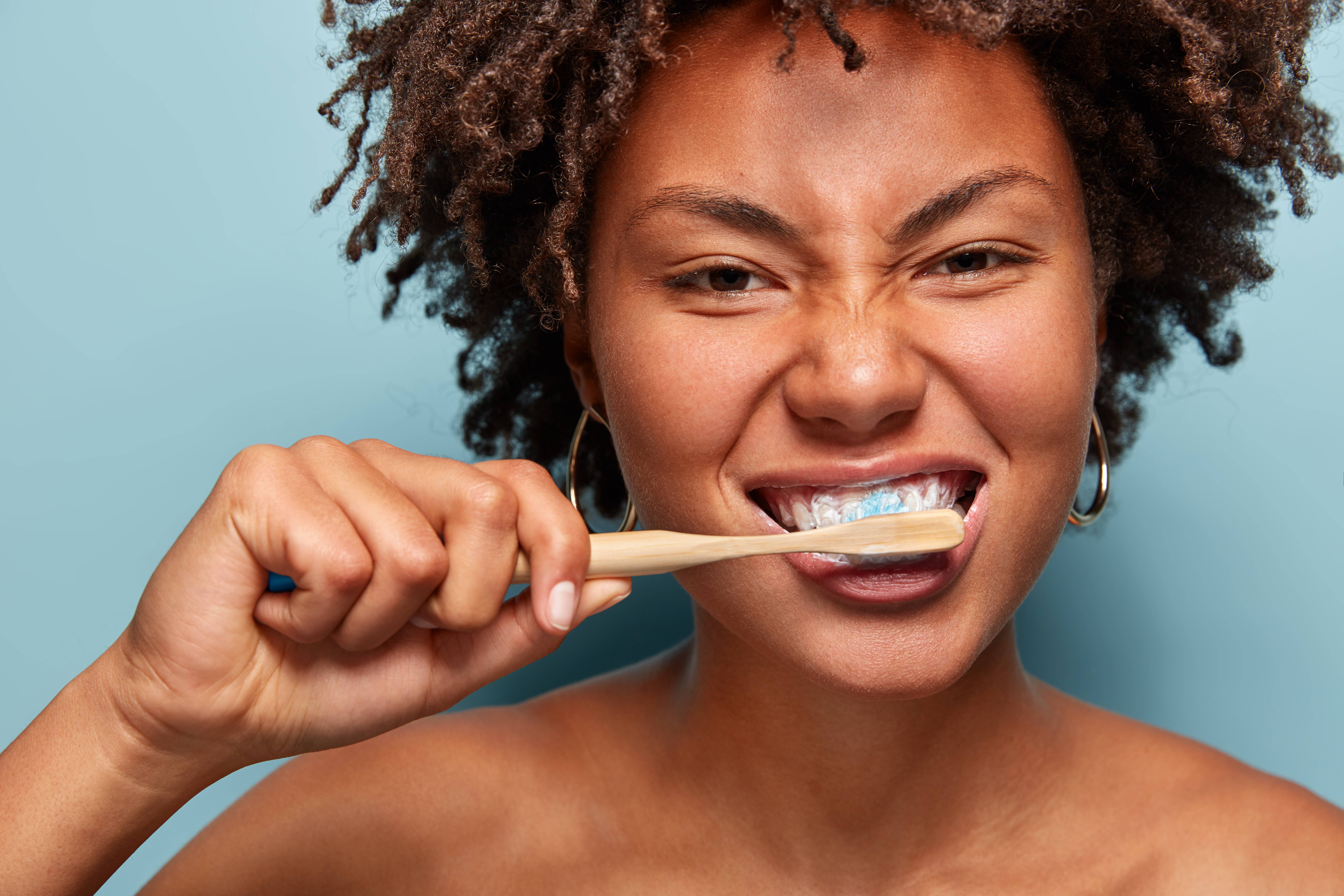 Dental hygiene concept. Self confident mixed race young woman brushes teeth with toothpaste and toothbrush, stands in front of mirror nude, isolated on blue background. Medicine, health care